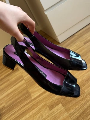 Svarta lackpumps från Betty Blue - Säljer ett par svarta lackpumps från Betty Blue med öppen tå och låg klack. Skorna har en slingback-rem och lila innersula i mjukt material.