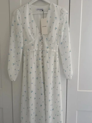 Vit blommig klänning - Vit klänning från adoore med blåa blommor.Helt ny, aldrig använd. Kom med en konstig doft men borde föravinna om man tvättar den en gång. Förmodligen någon fabrik/paketerings doft.