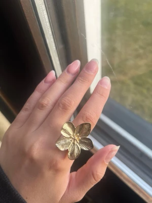 Stor guldfärgad blomring - Säljer en statementring i guldfärg med en stor, detaljerad blomma som motiv. Ringen har justerbar storlek och är tillverkad i metall. Perfekt för dig som vill ha en unik accessoar som sticker ut.