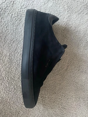 Svarta sneakers från Axel Arigato - Stilrena svarta sneakers från Axel Arigato i mocka med svart sula och diskret logga på sidan. Klassisk låg modell med minimalistisk design och rundad tå. Perfekta för dig som gillar en clean look och vill ha något som funkar till det mesta.