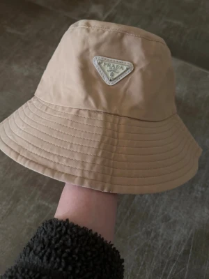 Beige bucket hat från Prada - Stilren beige bucket hat från Prada med ikonisk metalltriangel-logga framtill.  Använd max 1 gäng