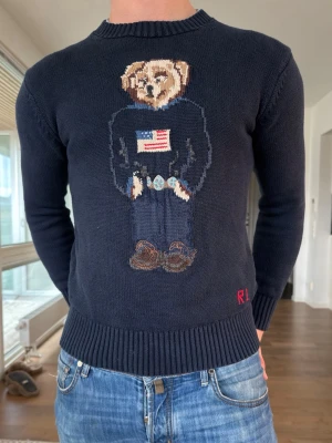 Polo Ralph Lauren stickad tröja - Säljer en superfin stickad tröja från Polo Ralph Lauren i klassisk marinblå färg (ser lite mörkare ut på bild). Tröjan har det ikoniska björn motivet framtill, vilket verkligen gör den unik och snygg  Storlek: M (sitter mer som en S) Skick: Väldigt fint skick, knappt använd Passform: Superbra – sitter riktigt snyggt på kroppen Färg: Marinblå  Perfekt till både vardag och lite mer uppklädda outfits.  Hör gärna av dig vid frågor och funderingar! 😊