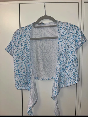 Blommig omlottopp med knytband - Söt kortärmad omlottopp med blått blommönster på vit botten. Toppen har knytband framtill och är croppad i modellen, perfekt för en somrig look. Materialet känns lätt och luftigt, vilket gör den skön att bära under varma dagar.