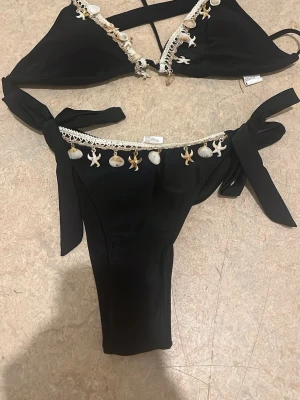 Svart bikini med snäckor och knyt - Snygg svart bikini med dekorativa snäckor och små sjöstjärnor längs kanterna. Både överdel och underdel har knytband i sidorna för justerbar passform. Perfekt för strandhäng och sommarens alla bad.