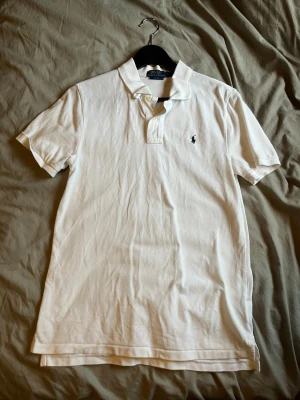 Vit pikétröja från Polo Ralph Lauren - Klassisk vit pikétröja från Polo Ralph Lauren i slim fit-modell. Tröjan har korta ärmar, krage med knappar och den ikoniska broderade loggan på bröstet. Tillverkad i mjuk bomull som känns skön mot huden. Perfekt till jeans eller shorts.