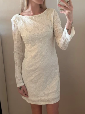 Vit spetsklänning med öppen rygg - Vit kortklänning med blommigt spetsmönster och långärmad design. Klänningen har en elegant öppen rygg och en kroppsnära passform som framhäver siluetten. Perfekt för dig som vill ha en stilren och feminin look. (Stolen S men passar M) säljes då den är för stor för mig!💕