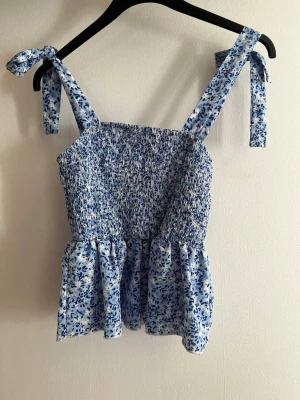 Blommig blå topp med knytband - Söt blå och vit topp med blommigt mönster och smockad överdel. Axelbanden knyts på axlarna för en personlig touch och toppen har en volang nedtill. Perfekt för varma dagar och ger en fräsch sommarkänsla. Ej använd