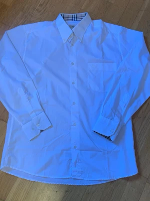 Vit skjorta från Burberry  - Klassisk vit skjorta från Burberry London med lång ärm och knappar framtill. Kragen har den ikoniska Burberry-rutiga detaljen på insidan. Skjortan har bröstficka och är tillverkad i bomull för en skön och stilren känsla.