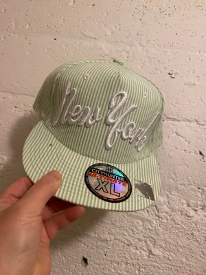 Randig New York keps från City Hunter - Säljer två snapback-kepsar från City Hunter med broderad 'New York'-text framtill. En keps är ljusgrön och vitrandig, den andra röd och vitrandig. Båda har platt skärm och klassisk streetwear-look. Storlek XL. Perfekt för dig som vill sticka ut med coola detaljer. 135kr/st 