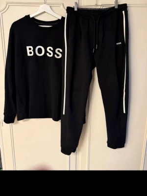 Svart BOSS set med vita detaljer - Säljer ett svart set från BOSS med långärmad tröja och byxor. Tröjan har stor vit BOSS-logga framtill och byxorna har vita ränder längs benen samt liten logga på fickan. Setet har en sportig och avslappnad look, perfekt för chill dagar. Materialet känns mjukt och bekvämt.