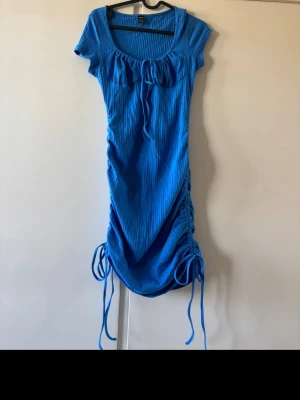 Blå ribbad kortklänning med snörning - Snygg blå ribbad klänning med kort ärm och snörningsdetaljer i sidorna samt framtill vid bysten. Klänningen har en tight passform och är tillverkad i ett stretchigt material som sitter skönt på kroppen. Perfekt för dig som gillar färg och trendiga detaljer.
