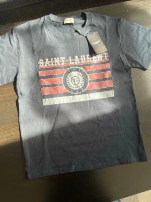 Saint Laurent t-shirt med tryck - Snygg svart t-shirt från Saint Laurent med coolt tryck framtill i rött, vitt och silver. T-shirten har rund halsringning och klassisk passform. Perfekt för dig som vill ha en statement tee från ett ikoniskt märke.