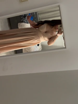 Lång ljusbeige klänning med omlott - Säljer en elegant långklänning i ljusbeige ton med omlottdetalj över bysten och breda axelband. Klänningen har ett flytande fall och markerad midja, perfekt för speciella tillfällen. Materialet känns mjukt och följsamt.