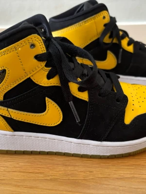 Nike Air Jordan 1 Mid svart/gul 37,5 - Snygga Nike Air Jordan 1 Mid sneakers i svart och gult. Skorna har klassisk hög siluett, svart mocka och gult läder med ikoniska swoosh-loggan på sidan. Tjock vit sula och svarta skosnören. Perfekta för dig som vill sticka ut med stilrena färger.