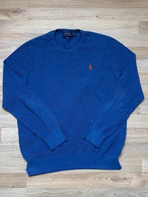 Blå tröja från Polo Ralph Lauren - Klassisk blå tröja från Polo Ralph Lauren med rund halsringning och den ikoniska logotypen broderad på bröstet. Tröjan har långa ärmar och ribbade muddar vid ärmslut och nederkant. Perfekt för en stilren och avslappnad look.
