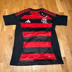 Flamengo 25/26 home kit L.Paqueta #20 - Säljer en svart och röd randig Flamengo matchtröja från Adidas med klubbmärke och sponsorlogga på bröstet. Tröjan har korta ärmar, rund hals och namn samt nummer 20 'L. Paquetá' på ryggen. Perfekt för fotbollsfans!