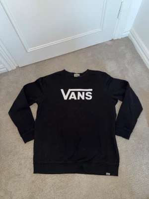 Svart tröja från Vans - Svart sweatshirt från Vans med klassisk vit logga framtill. Tröjan har rund halsringning, långa ärmar och ribbade muddar vid ärmslut och nederkant. Perfekt för en avslappnad och stilren look.