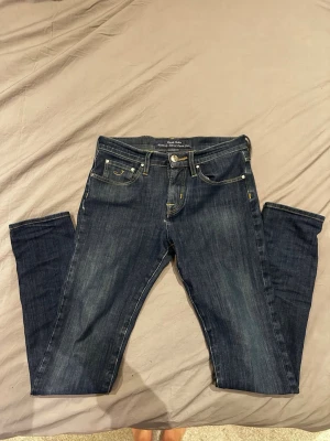 Mörkblå slim jeans från Jacob Cohen - Säljer ett par mörkblå jeans från Jacob Cohen med slim passform och klassisk femficksdesign. Jeansen har snygga slitningar framtill, gulddetaljer vid sömmarna och är tillverkade i mjukt denimtyg. Perfekta för dig som gillar stilrena och tidlösa jeans.