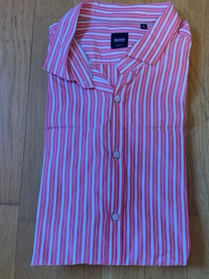 Rödvit randig skjorta Hugo Boss XL - Snygg röd och vit randig skjorta från Hugo Boss i slim fit-modell. Skjortan har klassisk krage, knäppning framtill och är tillverkad i ett tunt, skönt bomullstyg. Perfekt för dig som gillar stilrena och moderna skjortor med lite edge.