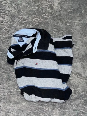 Tommy Hilfiger Striped Rugby Polo - Klassisk långärmad pikétröja (rugby-stil) i en snygg färgkombination med svarta, gråa och blåa ränder. Rejäl och skön kvalitet med snygga detaljer vid kragen.  • Skick: Mycket fint skick.