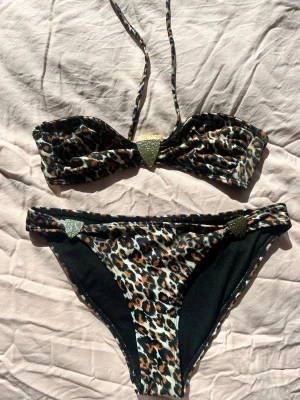 Leopardmönstrad bikini med gulddetaljer - Snygg bikini med leopardmönster och guldfärgade detaljer framtill på både överdel och underdel. Bandeau-topp med knyt runt nacken och matchande bikinitrosor. Aldrig använd, säljer då den inte passar 