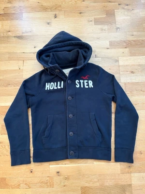 Vintage Hollister Cardigan med knappar - Säljer nu denna snygga marinblåa vintage Hollister cardigan. Tröjan har vit Hollister text broderat på framsidan, samt en vinröd Hollister logga. Står att tröjan är i storlek XL, men passar som L/M.  Skick 9/10, dvs i princip i nyskick. Modellen på bilden är 183 och väger ca 68 kilo.   Längd: 68 cm  Bredd (armhåla till armhåla): 58 cm  Skriv gärna om ni har några mer funderingar🙂