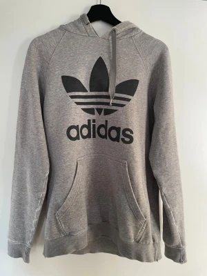 Adidas hoodie – grå med stor logga | M - Snygg grå hoodie från Adidas med stor klassisk trefoil-logga på bröstet 🔥  Storlek M (normal i storleken). Mjuk och bekväm med luva, snören och magficka – perfekt till vardag eller chill.  Använd men i bra skick, finns lite tecken på användning (se bild).