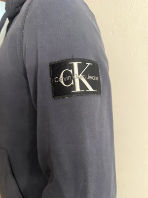 Mörkblå hoodie från Calvin Klein Jeans - Snygg mörkblå hoodie från Calvin Klein Jeans med stor CK-logga på ärmen. Hoodien har luva, magficka och ribbade muddar vid ärmslut och nederkant. Tillverkad i mjukt bomullsmaterial som är skönt att bära.