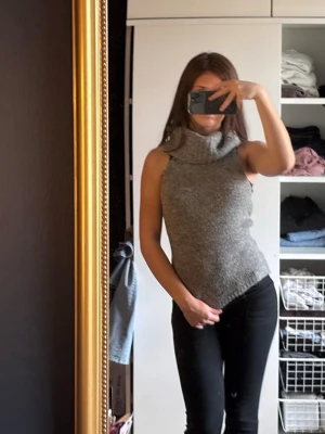 Grå stickad Gina topp  - Säljer en grå stickad topp med bred krage som kan bäras både som polokrage eller off-shoulder. Toppen är ärmlös och har en mjuk, följsam passform. Använd fåtal gånger 