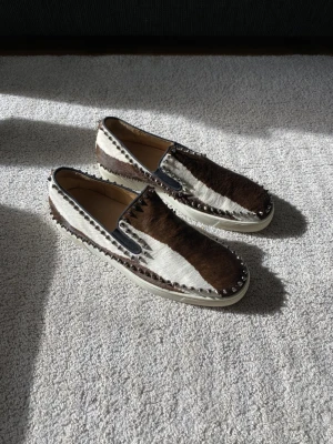 Christian Louboutin Roller Boat loafers med päls - Unika loafers från Christian Louboutin med brun och vit pälsimitation, dekorerade med nitar runt sulan. Skorna har den klassiska röda sulan och är tillverkade i Italien. Perfekta för dig som vill sticka ut med en exklusiv och edgy stil.
