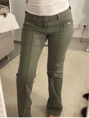 Olivgröna bootcut jeans med fickor - Snygga olivgröna jeans med bootcut passform och markerade sömmar framtill. Jeansen har två fickor med lock och knapp framtill samt bälteshällor. Materialet är stretchigt och bekvämt, perfekt för en trendig och avslappnad look.