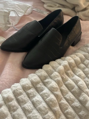 Svarta loafers med krokopräglat mönster - Svarta loafers med snygg krokopräglad yta och låg klack. Skorna har spetsig tå. Materialet ser ut som konstläder. Stora i storleken passar mig som brukar ha 38/39