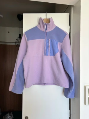 Ljuslila fleece med halv dragkedja - Mysig fleece i ljuslila och lavendelblått med hög krage och halv dragkedja framtill. Tröjan har en praktisk bröstficka med dragkedja och justerbar dragsko i nederkant. Perfekt för lager på lager och chill dagar.