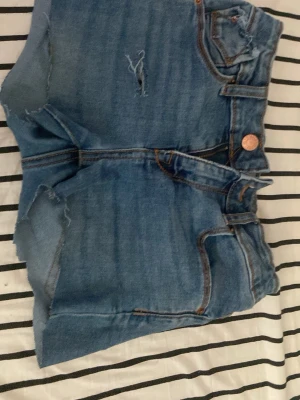 Blå denimshorts från 157 - Säljer ett par blå denimshorts från 157 med hög midja och råa kanter. Shortsen har klassiska fem fickor, slitningar framtill och kopparfärgade knappar. Perfekta för sommaren och ger en avslappnad vibe.och jag har gjort lite mer på dom