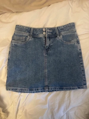 Blå jeanskjol med fickor - Snygg blå jeanskjol i klassisk modell med två framfickor, två bakfickor och bälteshällor. Kjolen har dubbla knappar och dragkedja framtill samt dekorativa sömmar. Perfekt för en trendig och avslappnad look.