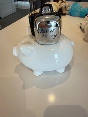 Spargris i keramik med resväska - Rolig spargris i vit keramik med en svart och silverfärgad resväska på ryggen. Perfekt för att spara till nästa äventyr eller resa. Spargrisen har klassisk form med små öron och nos, och väskan har gulddetaljer.