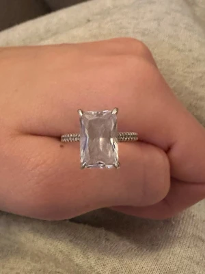 Silverring med stor rektangulär sten - Snygg silverfärgad ring med en stor, rektangulär klar sten i mitten. Ringen har en detaljrik banddesign med små räfflor längs sidorna. Perfekt för dig som gillar statement-smycken och vill sticka ut lite extra. Märke och material på stenen är okänt.