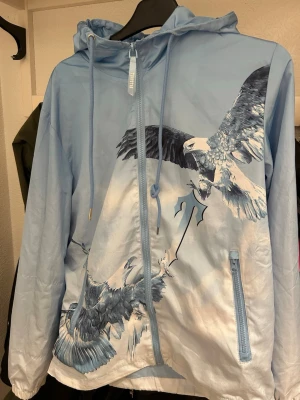 Ljusblå vindjacka med örntryck - Säljer en ljusblå Trapstar Eagle! Jackan har ett stort, detaljerat tryck av örnar både fram och bak samt snörning i huvan som har stor efterfrågan. Materialet är lätt och vindtätt, perfekt för blåsiga dagar. Jackan passar i storlek 165-174 