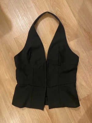 Svart halterneck korsettopp - Snygg svart korsettopp med halterneck och djup v-ringning. Toppen har en figurnära passform och markerad midja som ger en stilren siluett. Perfekt att styla med jeans eller kjol för en trendig look.