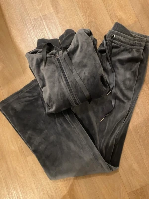 Mjuk mörkgrå velour loungewear-set - Säljer ett mysigt loungewear-set i mörkgrå velour. Setet består av en zip-hoodie med dragsko och matchande byxor med resår i midjan och snörning. Perfekt för chill dagar hemma eller när du vill vara extra bekväm.