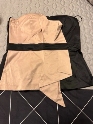 Beige och svart axelbandslös Buister  - Snygg axelbandslös buister i beige och svart med asymmetrisk detalj framtill. Buister har en markerad midja och är tillverkad i ett glansigt material som ger en lyxig känsla. Perfekt för dig som vill sticka ut med en unik look.