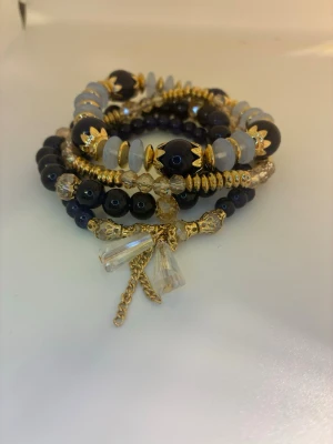 Flerfärgat pärlarmband med gulddetaljer - Snyggt armband med flera rader av pärlor i svart, blått och transparent, dekorerat med guldfärgade detaljer och kedjor. Armbandet har även hängande kristalliknande berlocker som ger extra bling. Ej använd utan kommer från min small business.