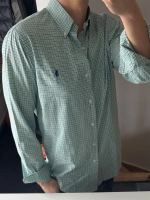 Grön rutig skjorta från Ralph Lauren - Klassisk grön och vit rutig skjorta från Ralph Lauren med blå logga broderad på bröstet. Skjortan har lång ärm, button-down krage och knappar framtill. Tillverkad i mjuk bomull och passar perfekt för en clean och stilren look.