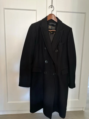 Svart dubbelknäppt kappa Zara Woman - Stilren svart kappa från Zara Woman med dubbelknäppning och klassisk krage. Tillverkad i samarbete med Manteco, känt för premiumtyger. Lång modell med fickor och snygga detaljer, perfekt för en trendig och elegant look.