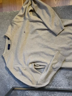 Beige tröja från GANT - Klassisk beige tröja från GANT med rund halsringning och diskret logga broderad på bröstet. Tröjan har långa ärmar och ribbade muddar vid ärmslut och nederkant. Perfekt för en stilren och enkel look.