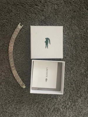 Lacoste silverfärgad armband - Säljer en silverfärgad klocklänk från Lacoste. Länken är i metall och har en klassisk design med rektangulära länkar. Perfekt om du vill byta ut eller ersätta din nuvarande klocklänk. Kommer i originalförpackning med Lacoste-logga.