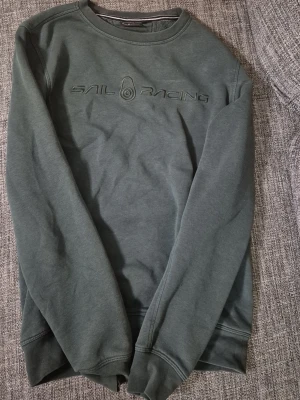 Mörkgrå sweatshirt från Sail Racing - Mörkgrå sweatshirt från Sail Racing med broderad logga på bröstet. Klassisk rund halsringning, ribbade muddar vid ärmslut och nederkant. Tillverkad i mjukt bomullsmaterial för en skön och avslappnad look. Passar i xs också