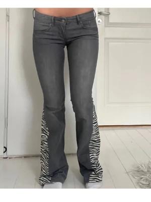 Lågmidjade gråa bootcut jeans med zebramönster - Säljer ett par gråa bootcut jeans med zebramönstrade paneler längst ner på benen. Jeansen har klassisk femficksmodell, låg midja och är i stretchigt material. Perfekta för dig som vill sticka ut med en unik detalj! Modellen är 159cm lång. Midjemått rakt över:38cm, innerbenlängd:80cm