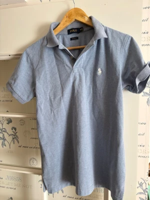 Ljusblå pikétröja från Polo Ralph Lauren - Snygg ljusblå pikétröja från Polo Ralph Lauren i modellen Custom Fit. Tröjan har korta ärmar, klassisk krage och den ikoniska vita broderade loggan på bröstet. Tillverkad i mjuk bomull som känns skön mot huden. Perfekt för en avslappnad men stilren look.