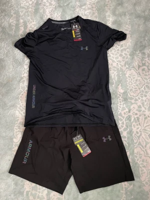 Svart träningsset från Under Armour - Snyggt svart träningsset från Under Armour med t-shirt och shorts. Båda plaggen har logga och text i regnbågsskimrande färger. Tillverkade i lätt och snabbtorkande material som passar perfekt för gym eller löpning.